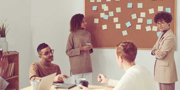 A group of men and women sit at a desk and stand at a cork board with post it notes on it